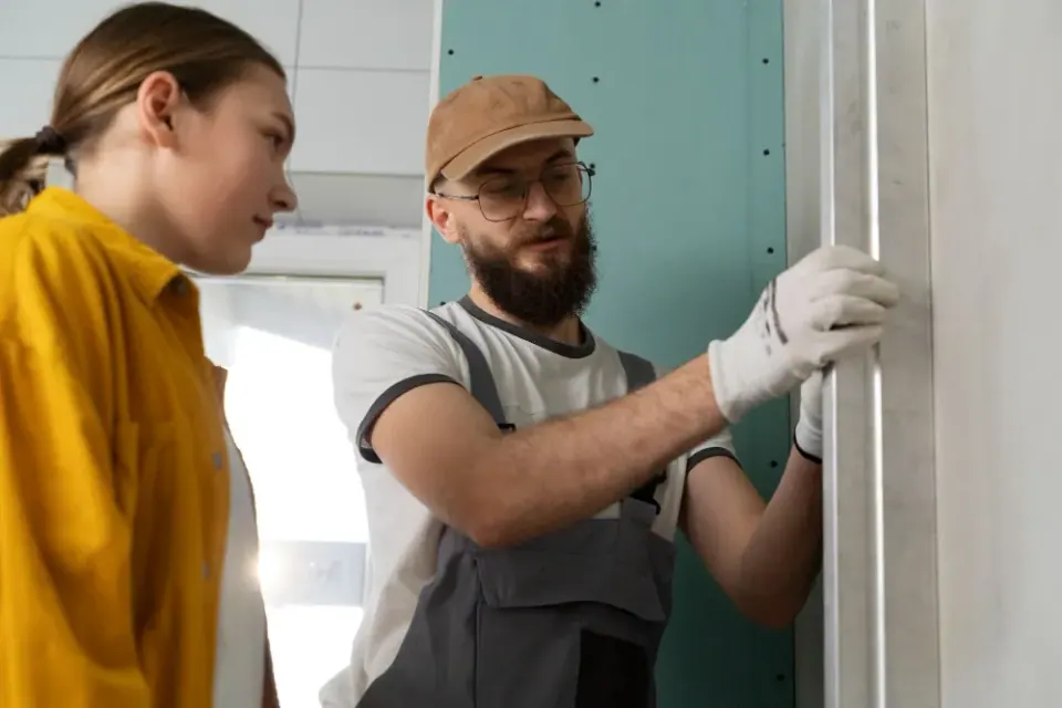 Man with beard, in work clothes, explains framing to a woman in yellow shirt; indoors, construction site.