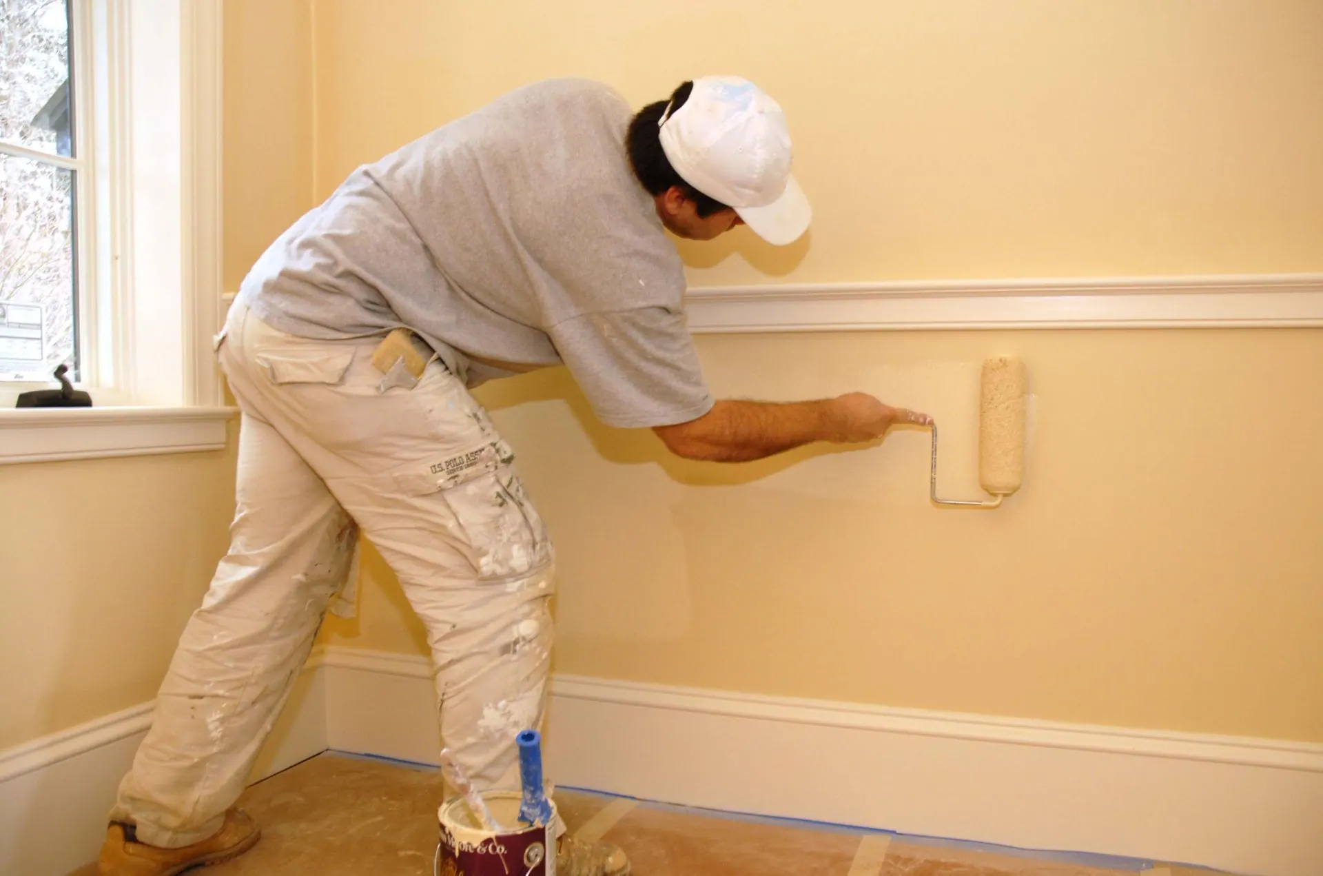 Person paints a wall with a roller. The wall and trim are beige. He wears a hat and work clothes.