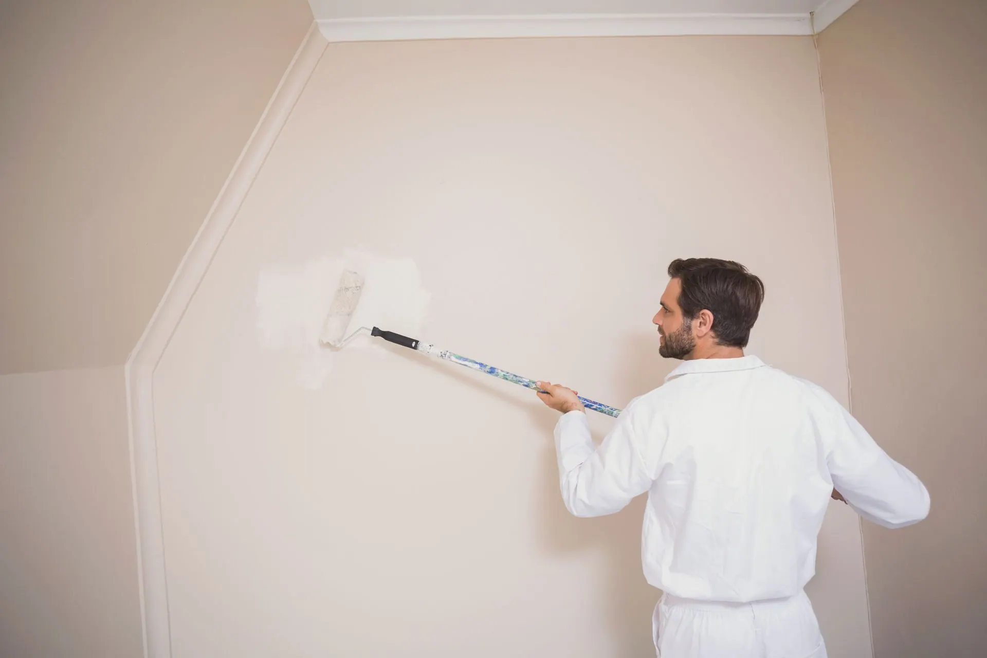 Man in white coveralls paints a wall with a roller. Beige walls and white trim in a room.